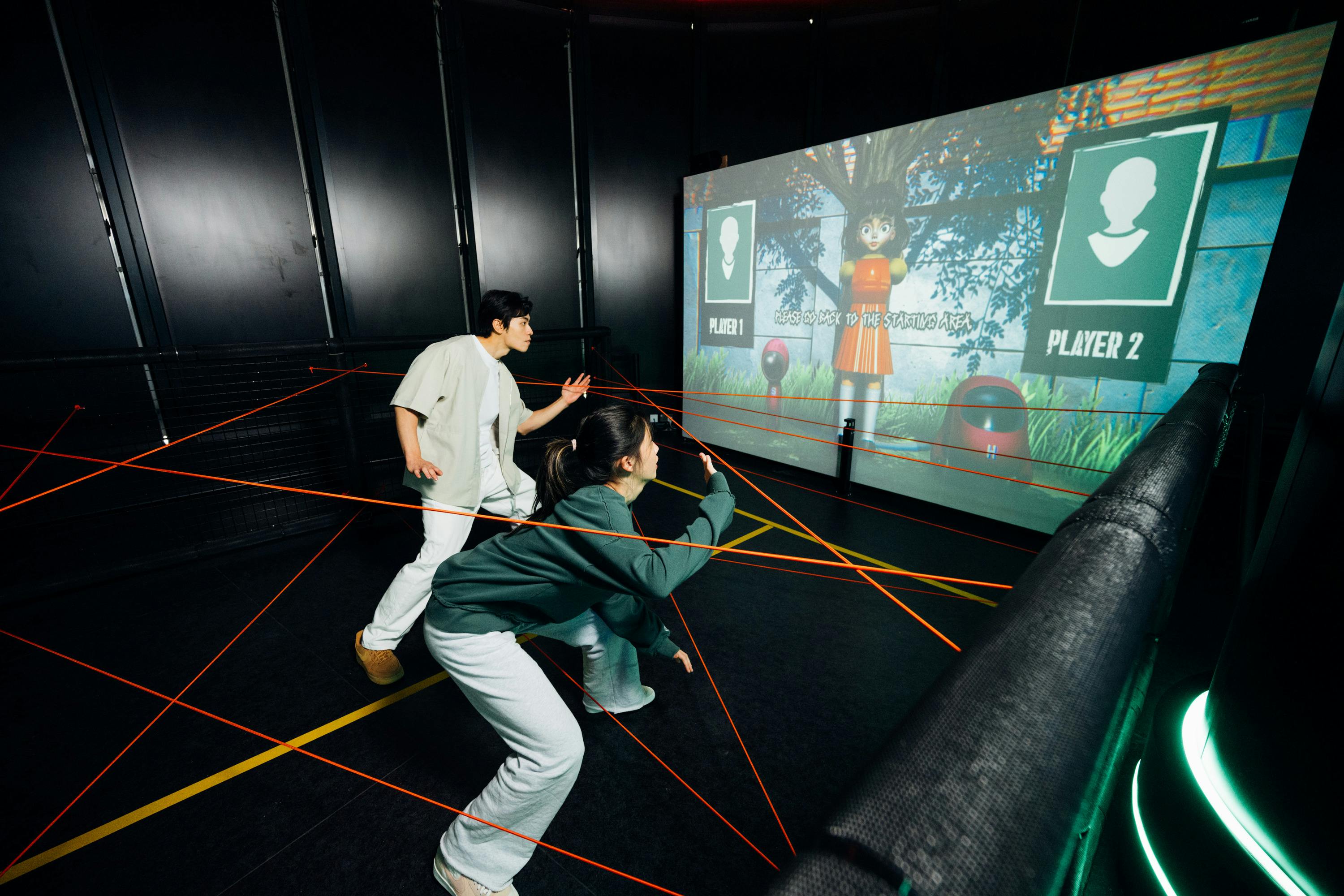 Two people navigate a room with red laser-like wires, focusing on a large screen showing a game with characters and instructions.