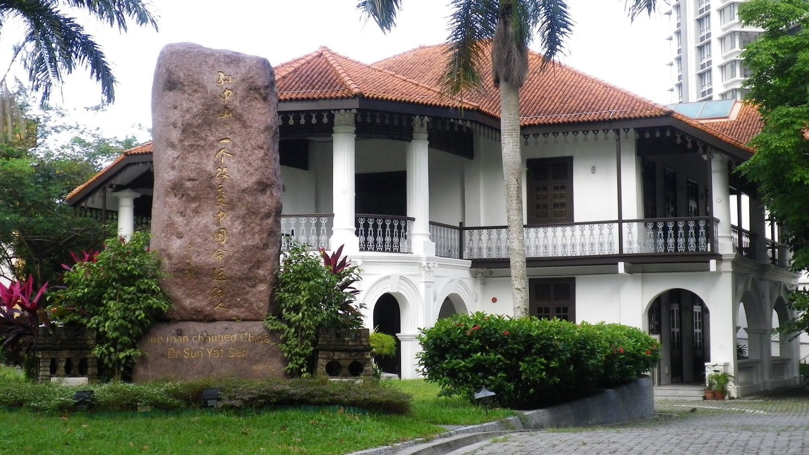 Un grande monumento in pietra con iscrizioni si trova di fronte a un edificio a due piani in stile coloniale circondato da una rigogliosa vegetazione.