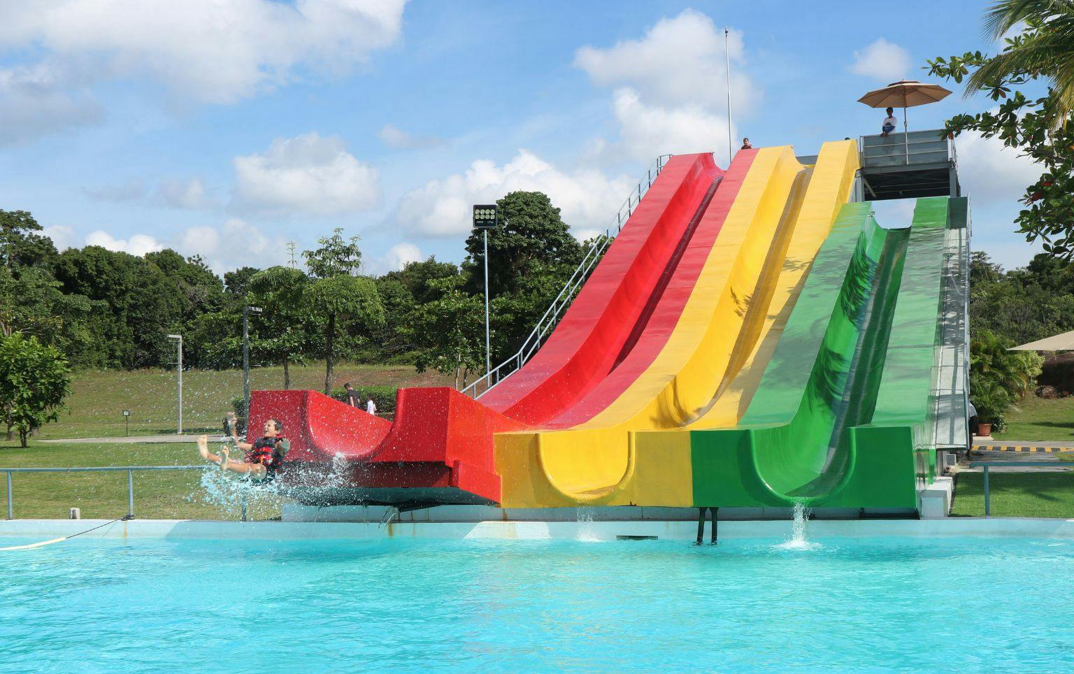A series of colorful water slides ending in a clear pool, surrounded by trees under a blue sky.