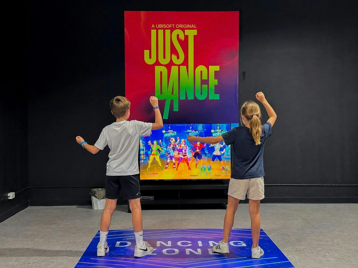 Two children playing a dance video game in front of a screen, with a "Just Dance" poster in the background.