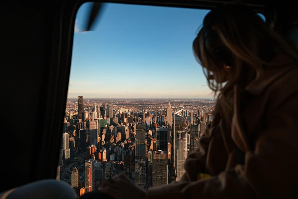 A person in a helicopter overlooking a city skyline during sunset with buildings and a clear sky visible.
