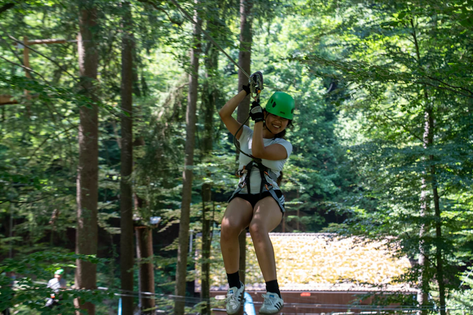 A person wearing a green helmet and safety harness ziplining through a forested area on a sunny day.