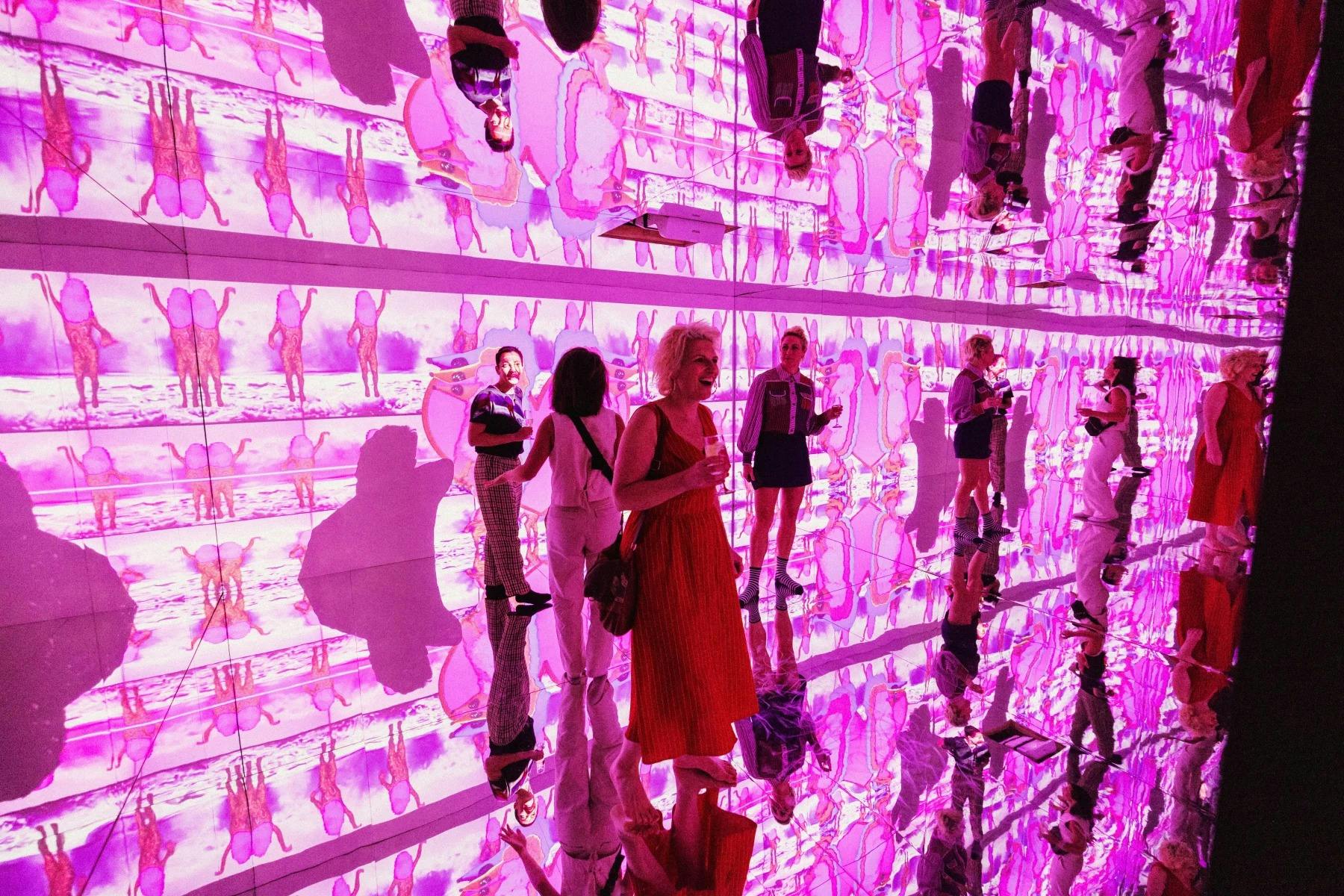 반사되는 바닥과 벽이 있는 방에 서 있는 사람들은 선명한 분홍색과 보라색 빛 투영으로 둘러싸여 있습니다.