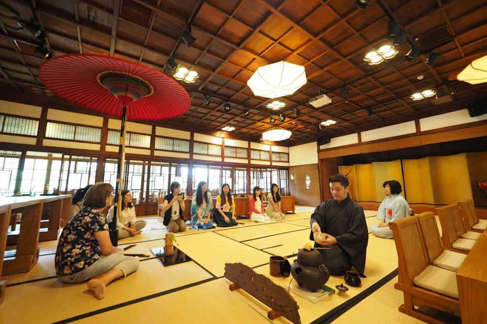 A traditional Japanese tea ceremony in a wooden room, with participants seated on tatami mats observing the host.