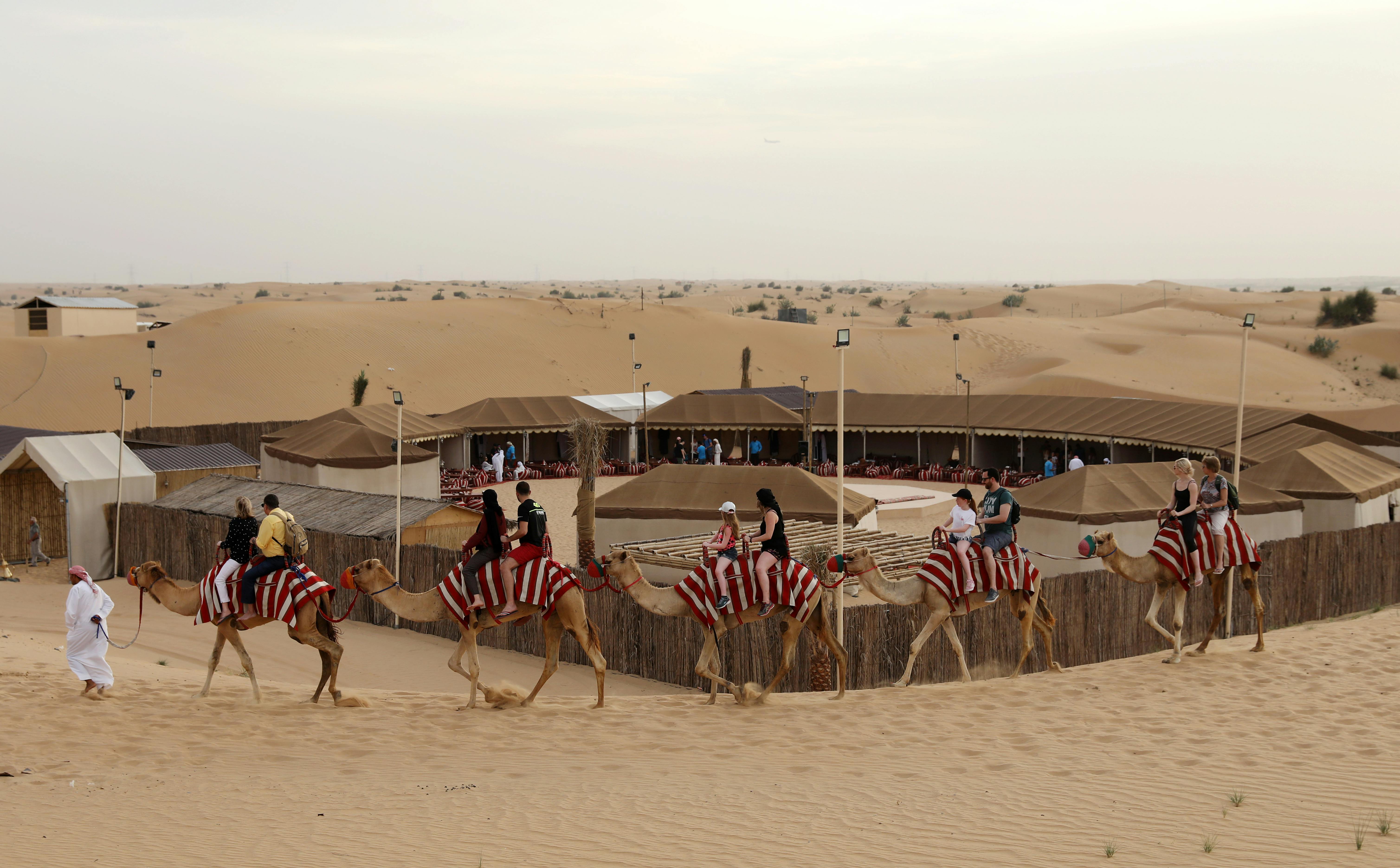 Lidé jezdící na velbloudech s červeně pruhovanými přikrývkami v poušti, míjející tábor se stany a dřevěnými ploty.