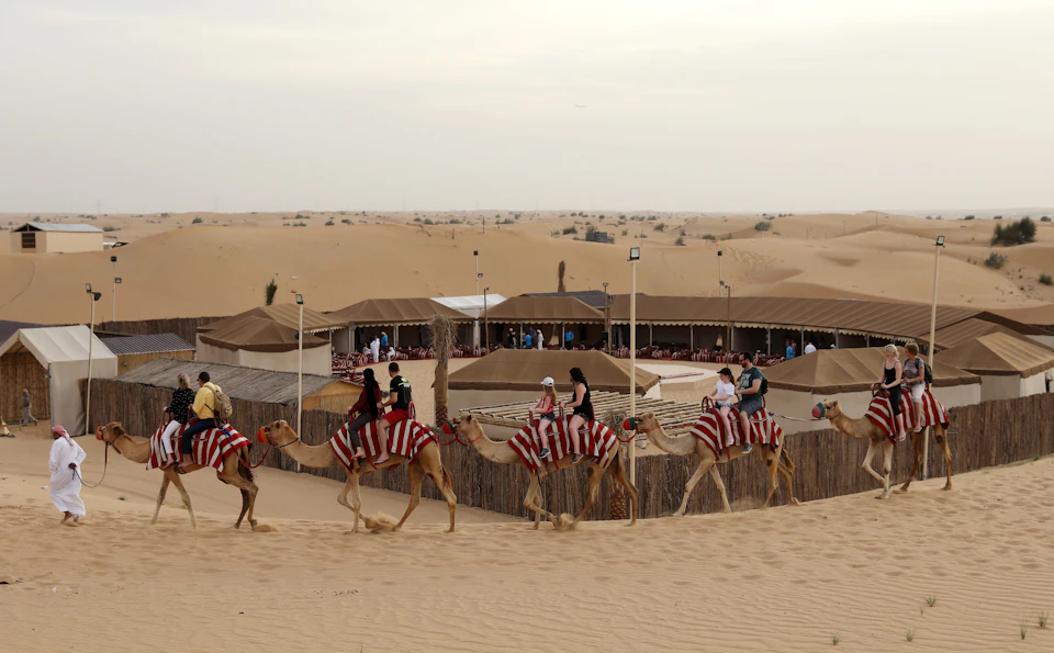 People riding camels with red-striped blankets in a desert, passing a camp with tents and wooden fences.