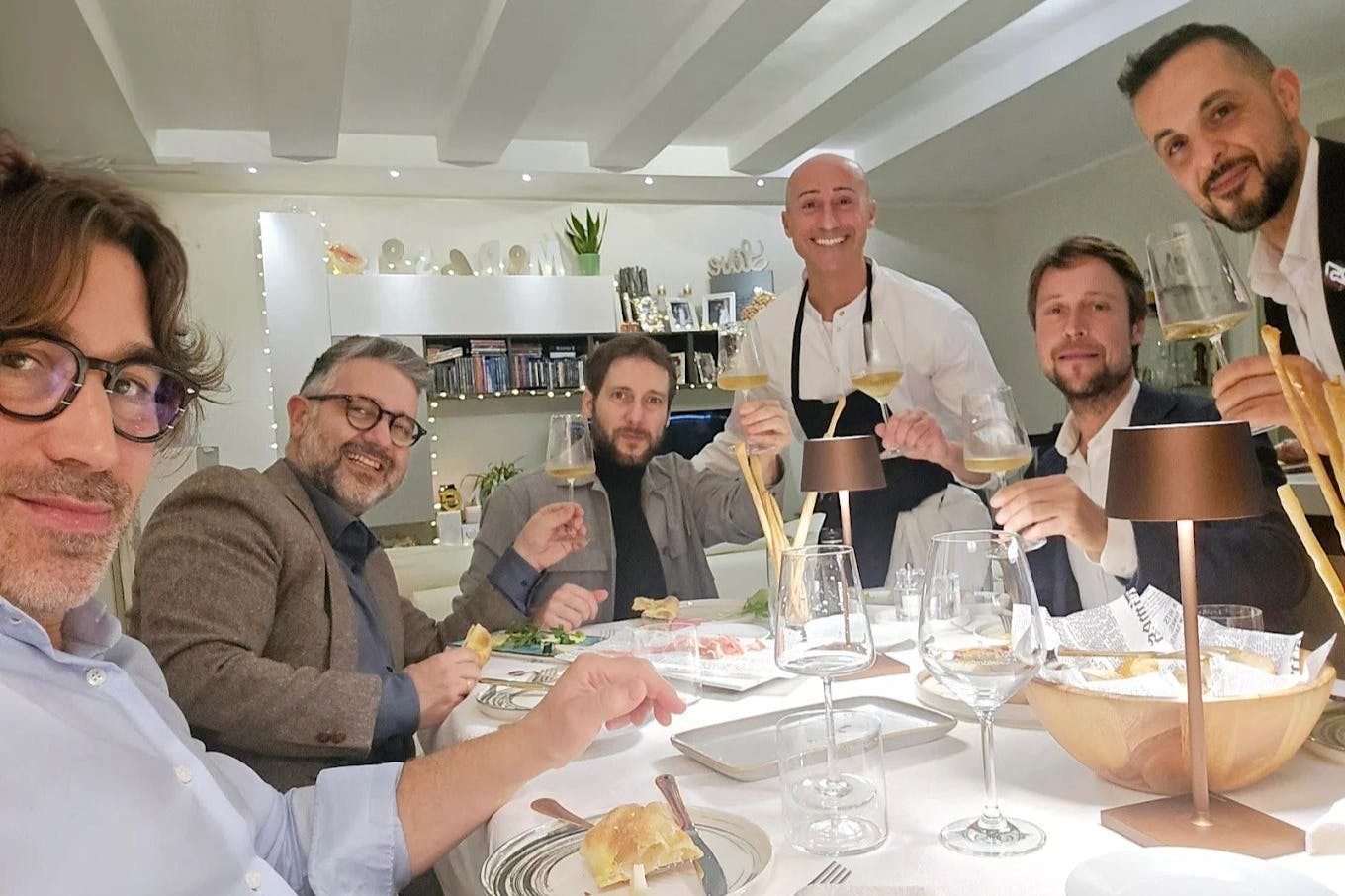 Four people seated at a dining table with a waiter, all holding glasses and smiling, in a cozy, well-lit dining room.