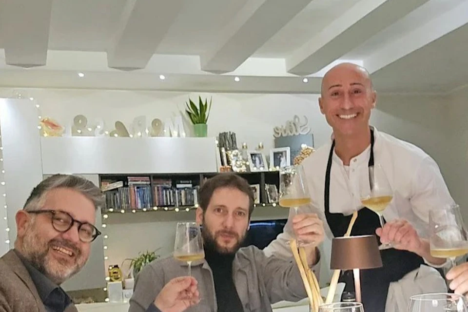 Four people seated at a dining table with a waiter, all holding glasses and smiling, in a cozy, well-lit dining room.