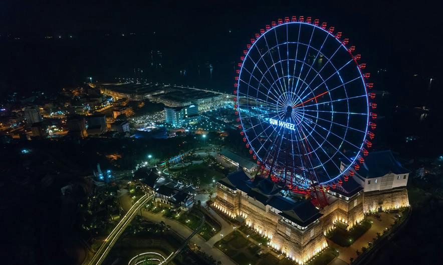 An illuminated Ferris wheel labeled 'Sun Wheel' and surrounding lit buildings at night in a cityscape.