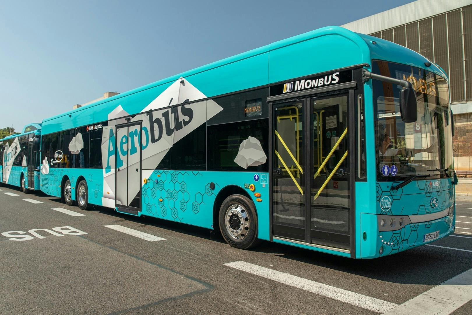 Un bus turquoise portant l'inscription "Aerobus" et "Monbus" sur le côté, garé sur une surface en béton à côté d'un bâtiment.