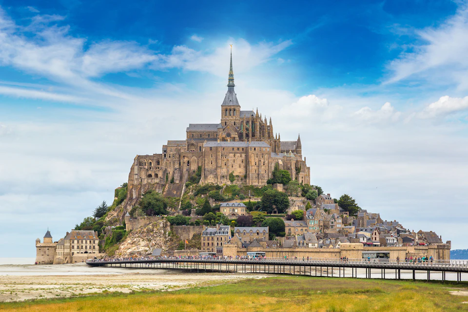 A historic abbey and village on a rocky island, connected by a bridge, with a bright blue sky and some clouds overhead.
