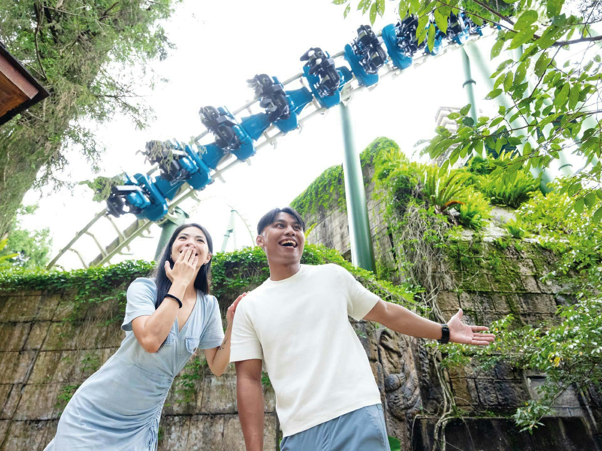 Two people standing outdoors smiling with a rollercoaster in the background surrounded by greenery.