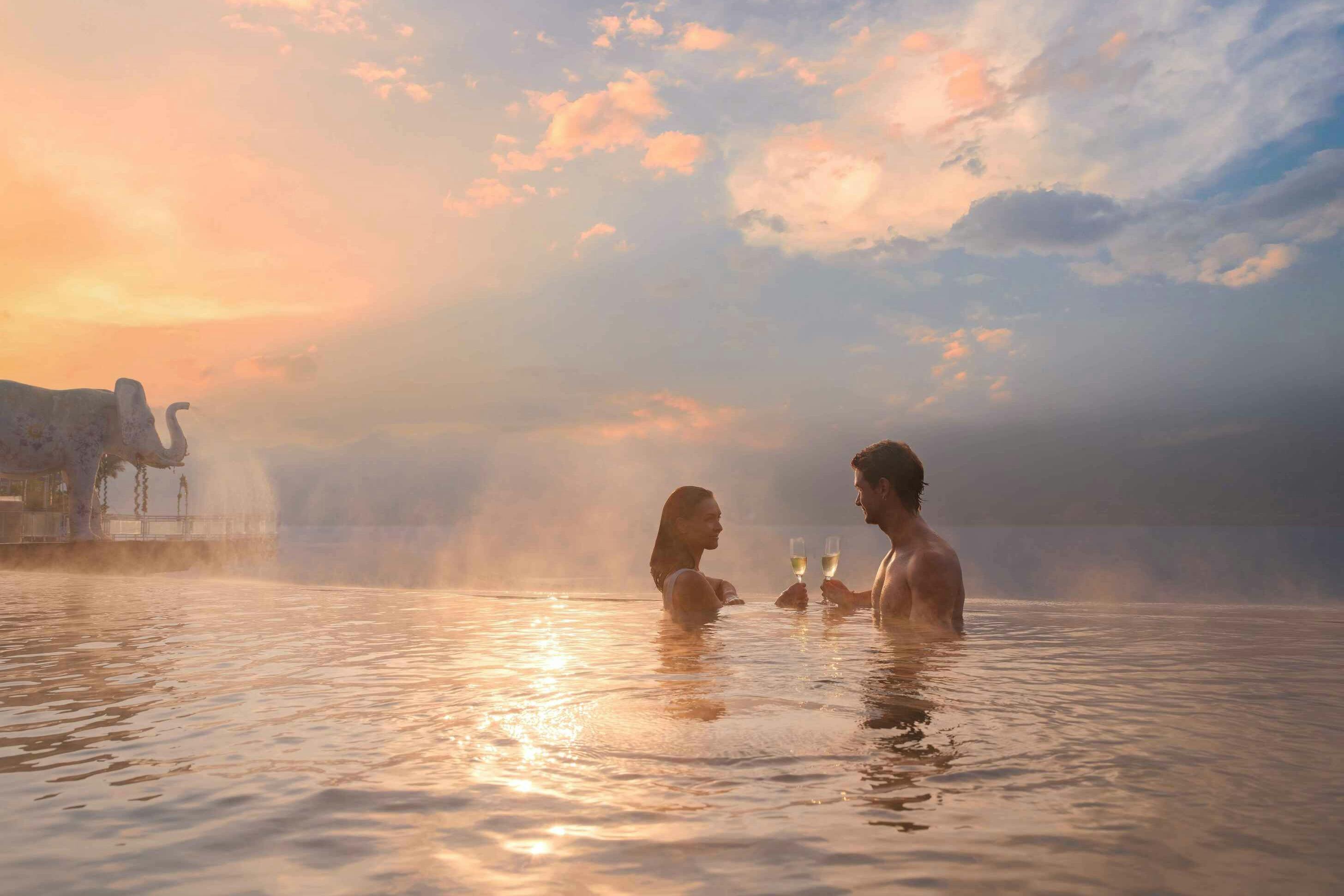 A couple standing in a serene, steaming pool at sunset, toasting with champagne glasses under a partly cloudy sky.