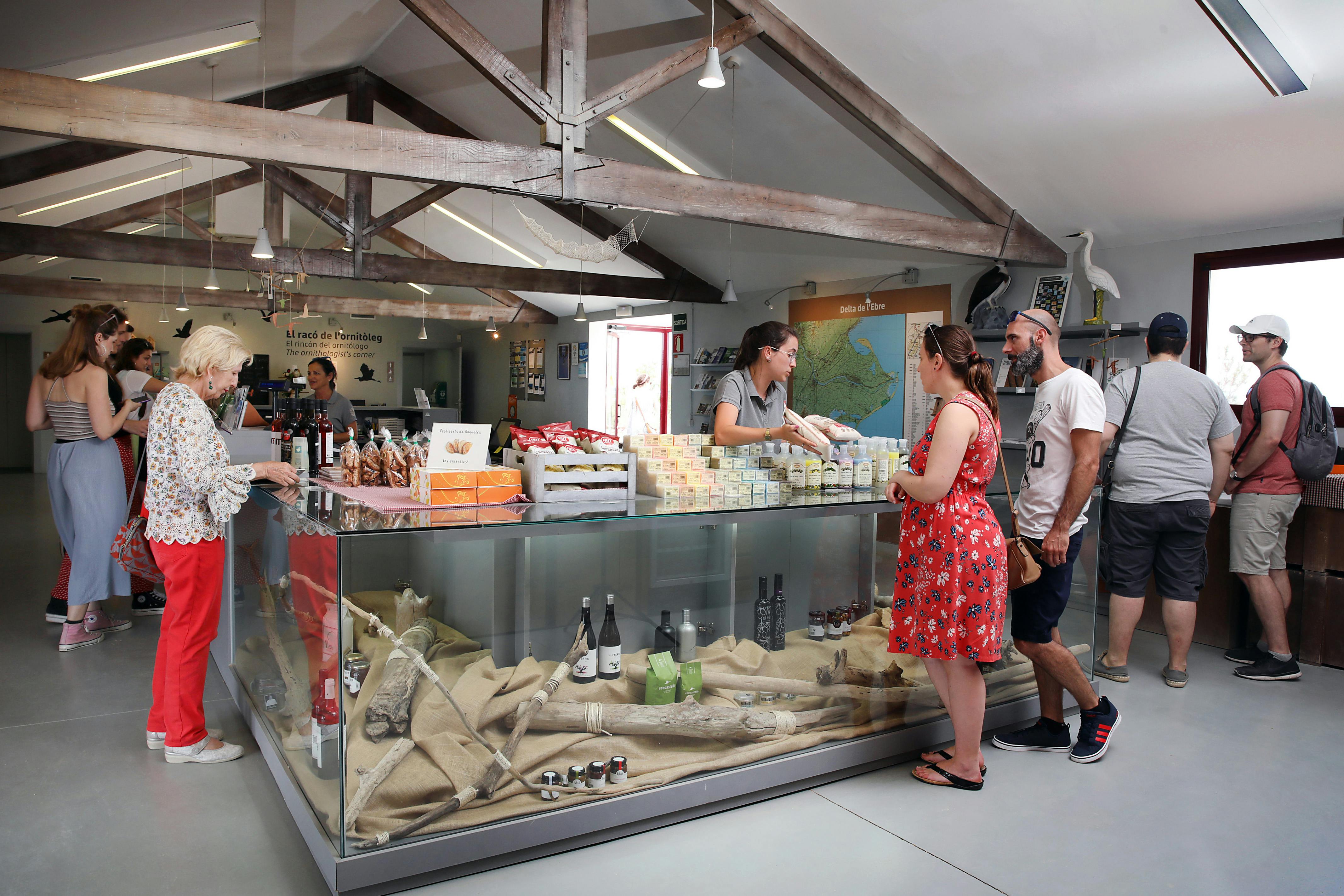 Lidé u dřevěné přepážky v obchodě, mluví s pokladníkem. Na regálech jsou vystaveny různé produkty, včetně krabicových předmětů a baleného zboží.