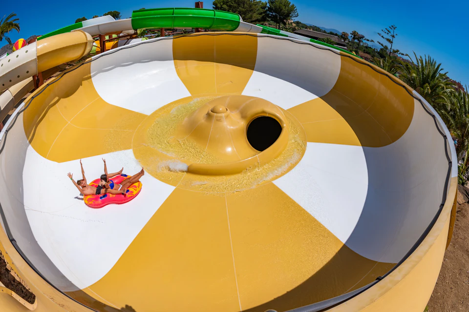 Two people in a raft sliding on a yellow and white water slide bowl at a water park, surrounded by trees and a clear sky.