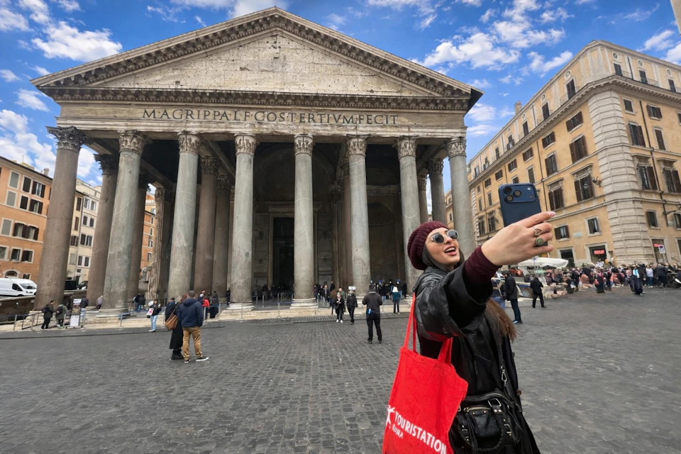 A person taking a selfie in front of the Pantheon in Rome, with a red tote bag and a clear sky above.