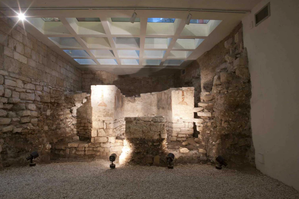 Ancient stone ruins are illuminated by spotlights under a glass skylight in a museum-like setting.