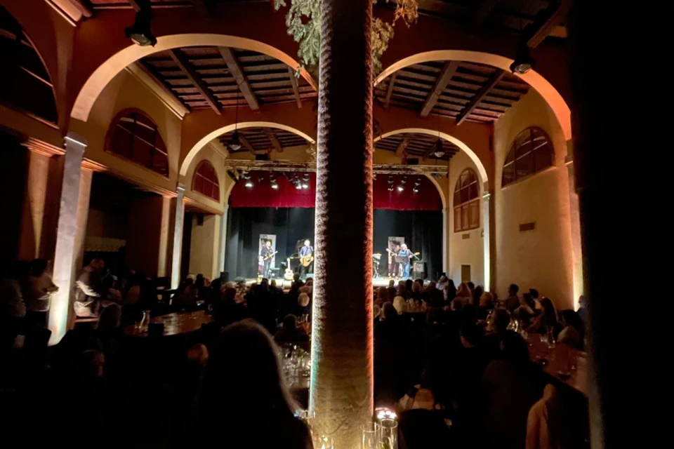 A band performs on a stage in a dimly lit venue with arched ceilings and a central pillar. An audience is seated, watching.