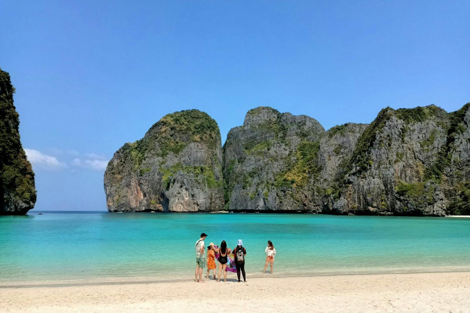 Skupina lidí stojí na písečné pláži s tyrkysovou vodou a velkými zelenými útesy v pozadí pod jasnou modrou oblohou.