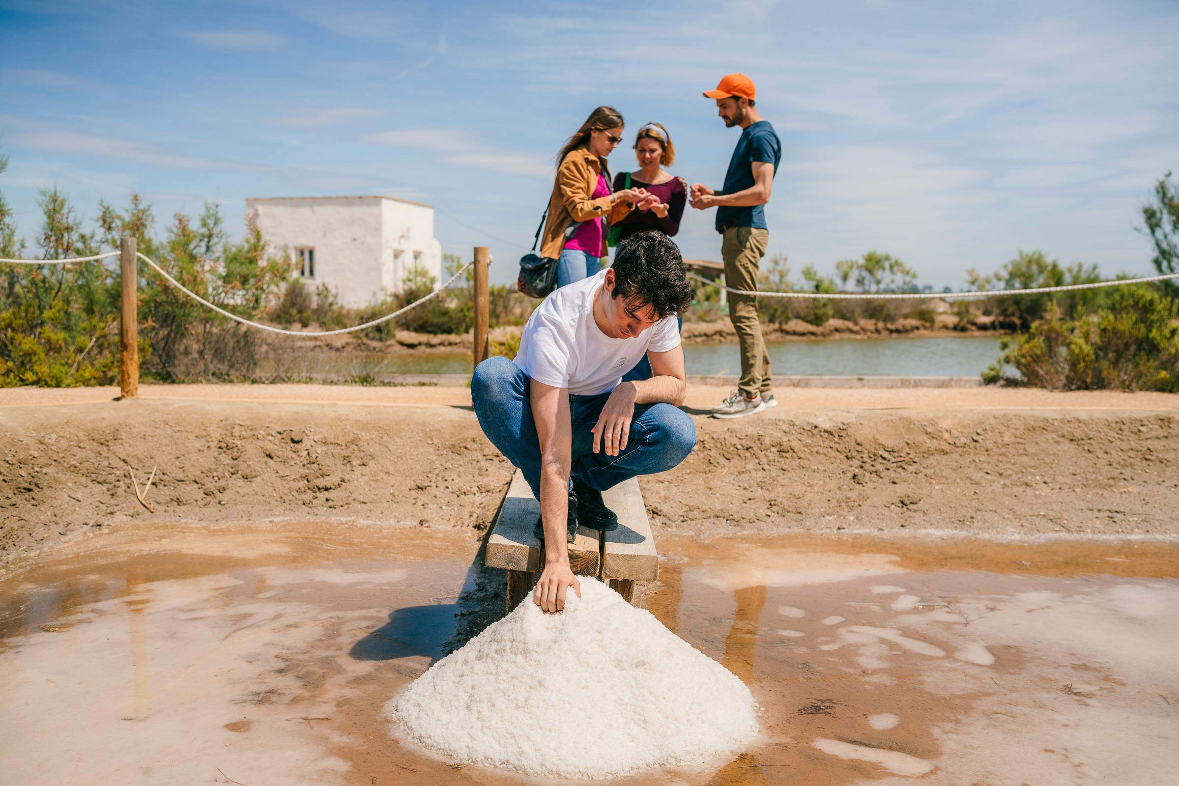 Muž nabírá sůl z hromady, zatímco tři lidé konverzují v pozadí poblíž bílé budovy a vody.