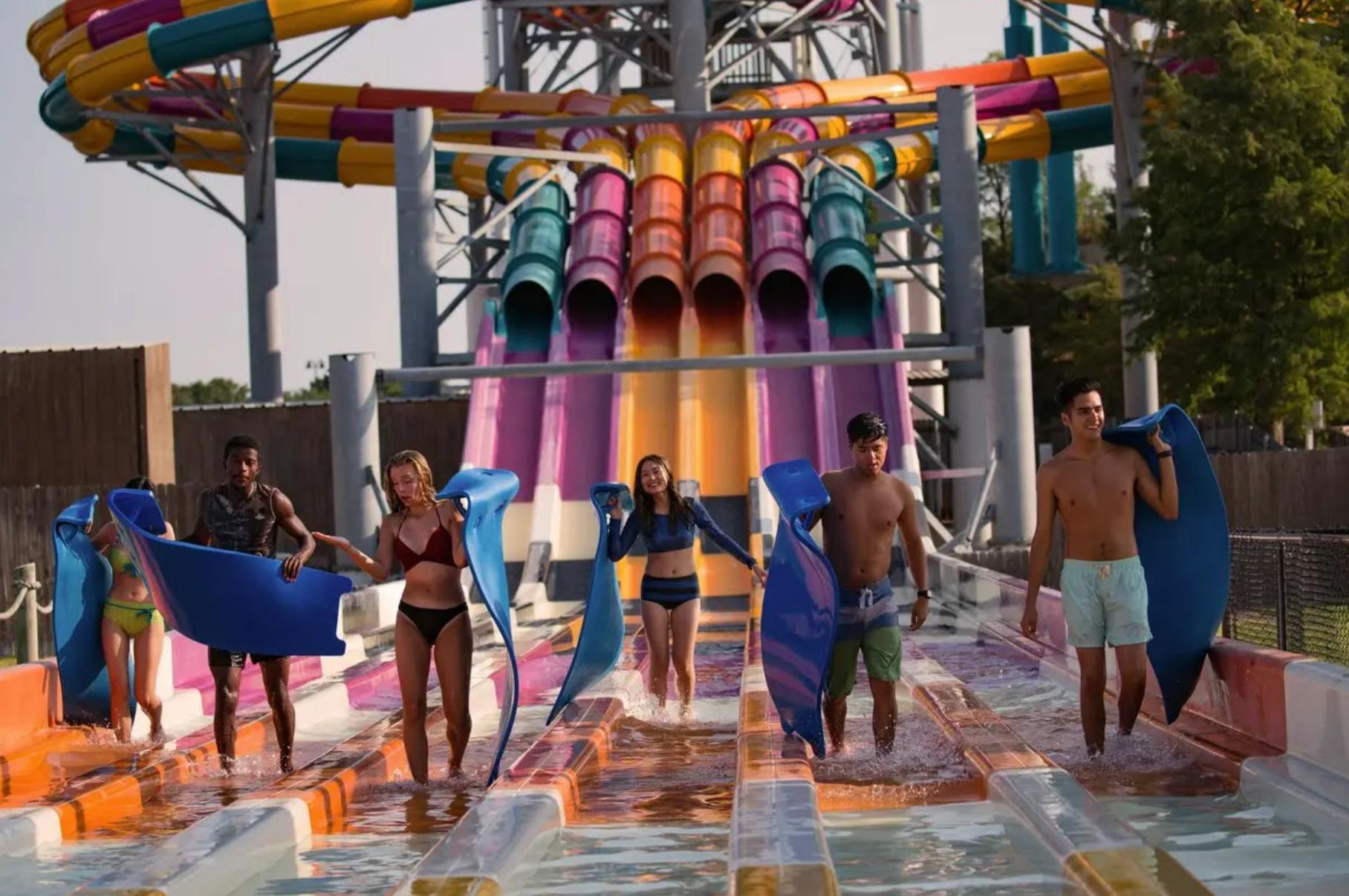 Quatre persones que sostenen estores caminen per aigües poc profundes al final de tobogans d'aigua de colors en un parc aquàtic a l'aire lliure.