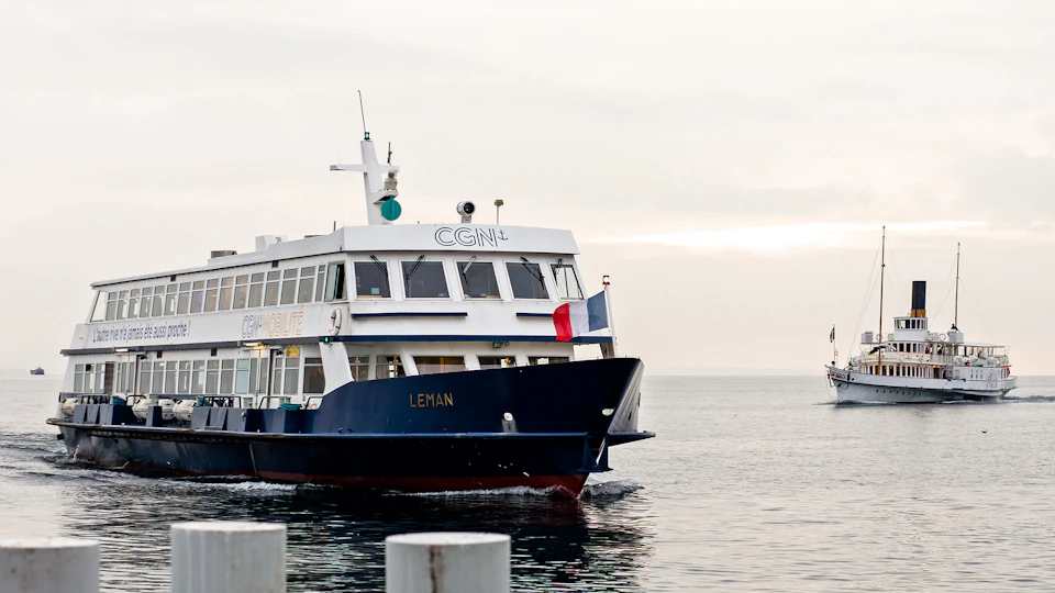 A ferry named "LEMAN" sailing on calm waters with a French flag at the front. Overcast sky in the background.