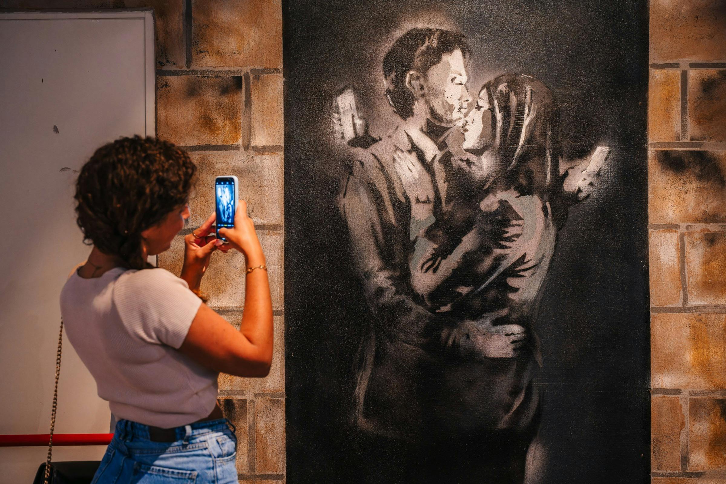 Une personne photographie une peinture murale représentant un couple enlacé, chacun tenant un téléphone, peinte sur un mur de briques.