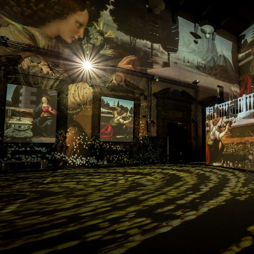 Room with large projections of classical paintings on walls, including images of a woman and detailed art scenes, under dim lighting.