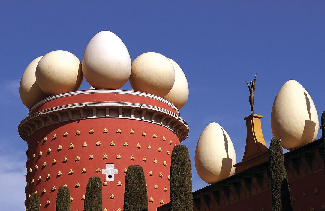 Un bâtiment rouge surmonté de grandes sculptures d'œufs et la statue d'une personne debout sur un toit, sous un ciel bleu clair.