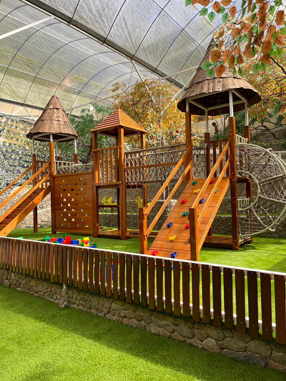 A children's playground with wooden structures, slides, climbing walls, and netting, surrounded by a grass area and fencing.