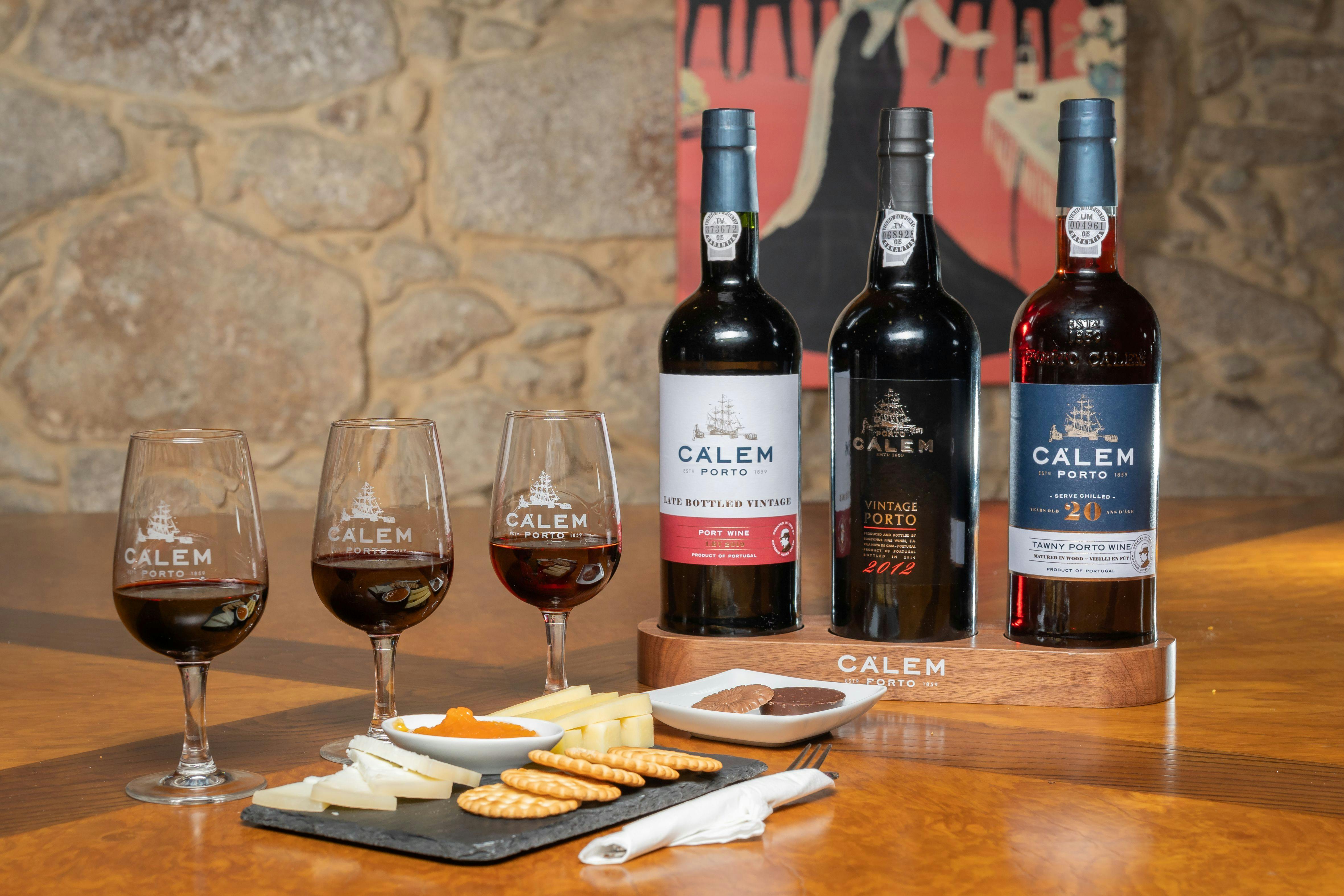 Three bottles of Calem Porto wine with three glasses of wine, cheese, and crackers arranged on a table in a rustic setting.