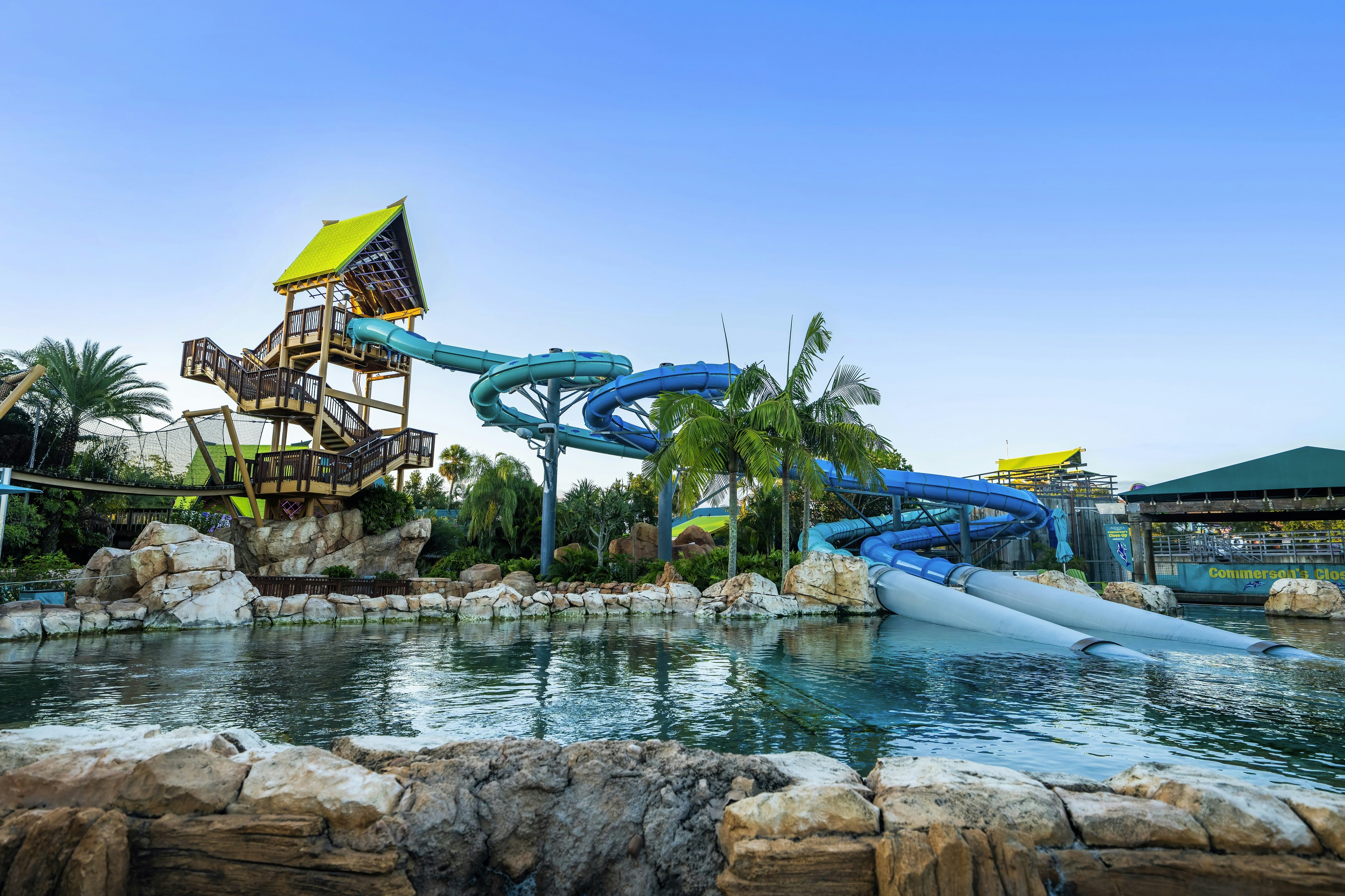 Ein tropischer Wasserpark mit einer großen, gewundenen blauen Wasserrutsche, Palmen, Felsen und einem Pool unter strahlend blauem Himmel.
