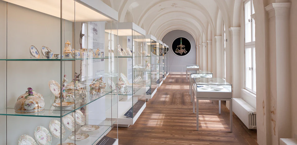 A museum corridor with glass display cases showcasing porcelain items, arches in the ceiling, and a chandelier at the end.