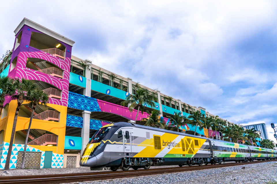 A modern Brightline train passes by a multicolored building with murals and palm trees under a cloudy sky.