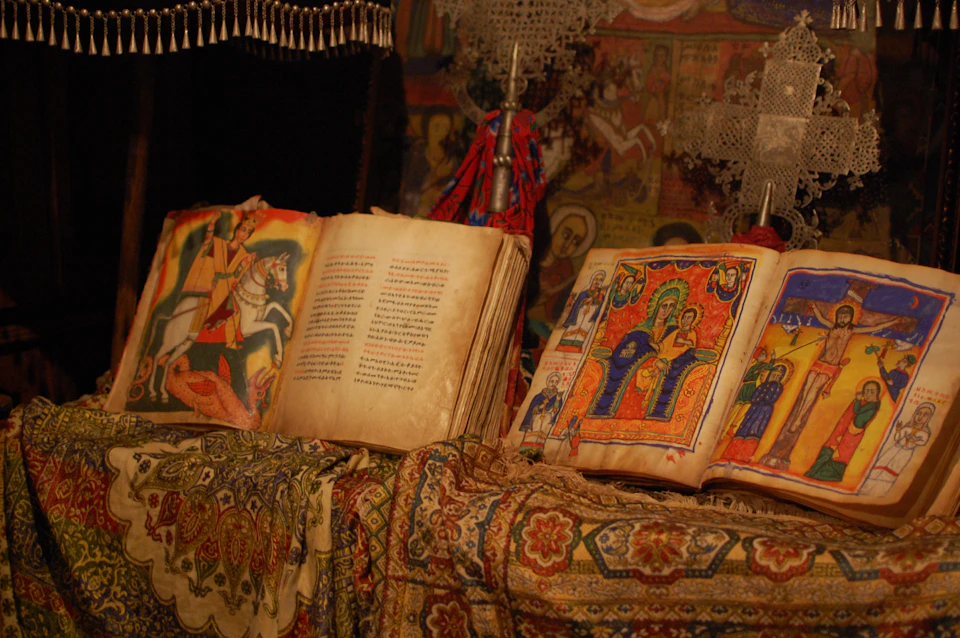 Old, ornate books with colorful illustrations are open on a richly patterned cloth, surrounded by religious artifacts.