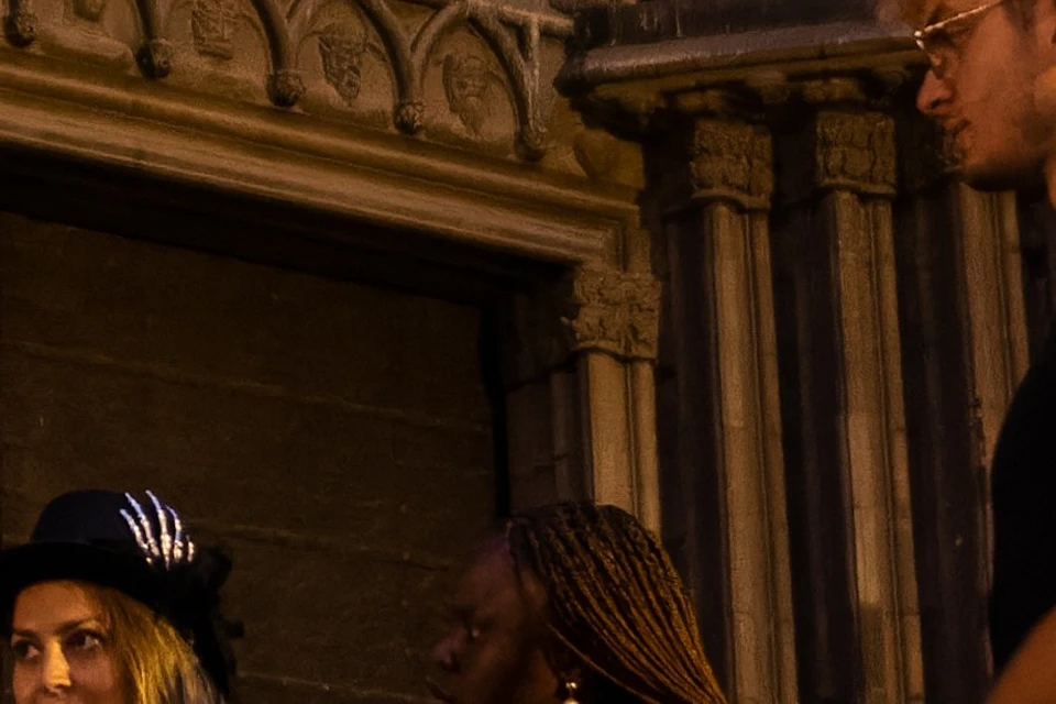 A woman in costume holding a lantern speaks to a group of people outside a historic building at night.