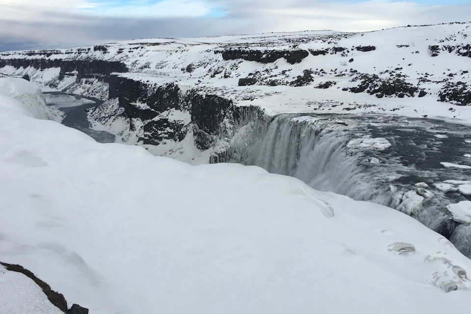 A large waterfall cascades over a cliff into a snowy landscape under a cloudy sky.