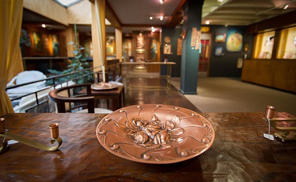 Wnętrze galerii sztuki z ozdobną miedzianą płytą na drewnianym stole na pierwszym planie i malowidłami na ścianach.