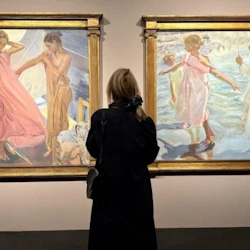 En person i en mörk kappa som observerar två inramade målningar med figurer i flytande plagg och utomhusscener.