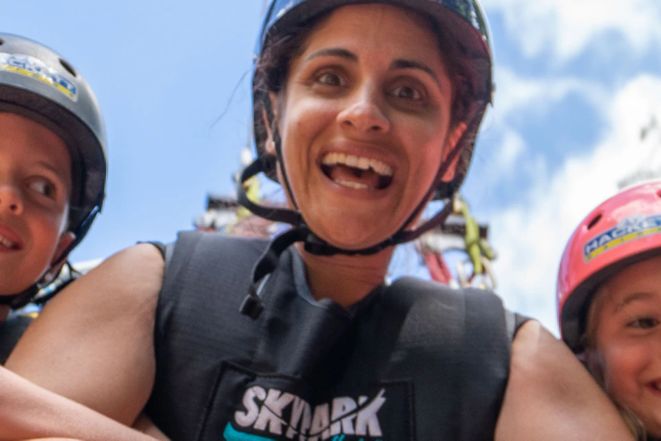 Three people, smiling and wearing harnesses and helmets, prepare for a zipline adventure against a backdrop of trees and sky.
