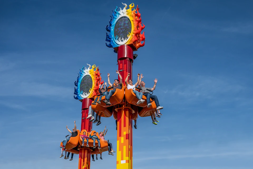 People sitting on colorful amusement park rides with flame-themed decorations, raising their hands, against a clear blue sky.
