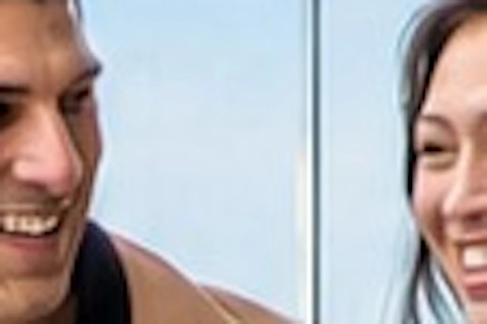 A man and woman walking on a glass-walled terrace, holding drinks, with a cityscape visible in the background.