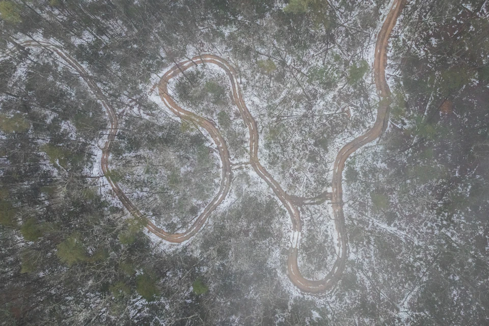 Aerial view of a winding dirt path through a snowy forest landscape with scattered trees.