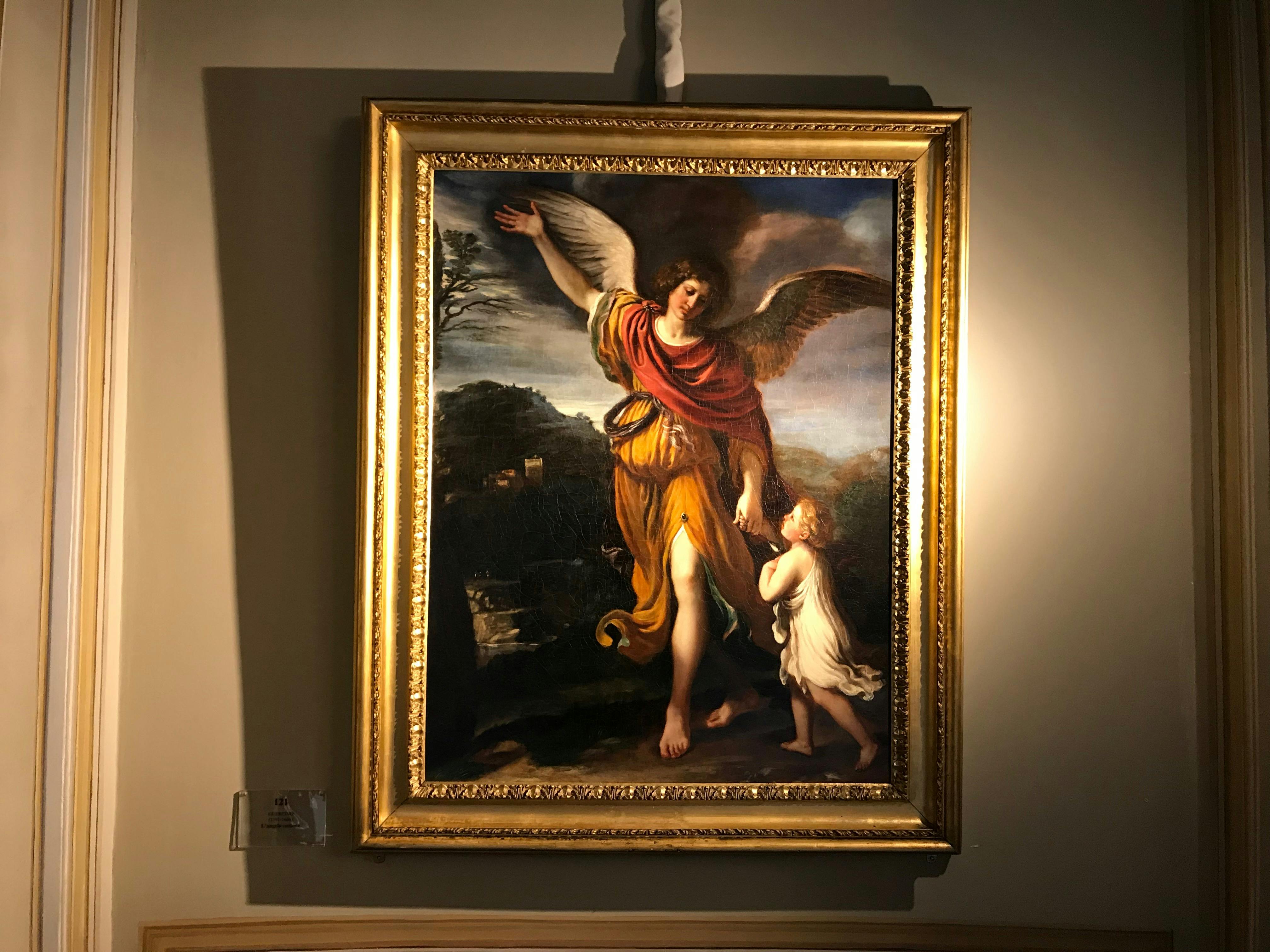 A framed painting depicting a winged figure guiding a smaller figure, illuminated in a gallery setting.