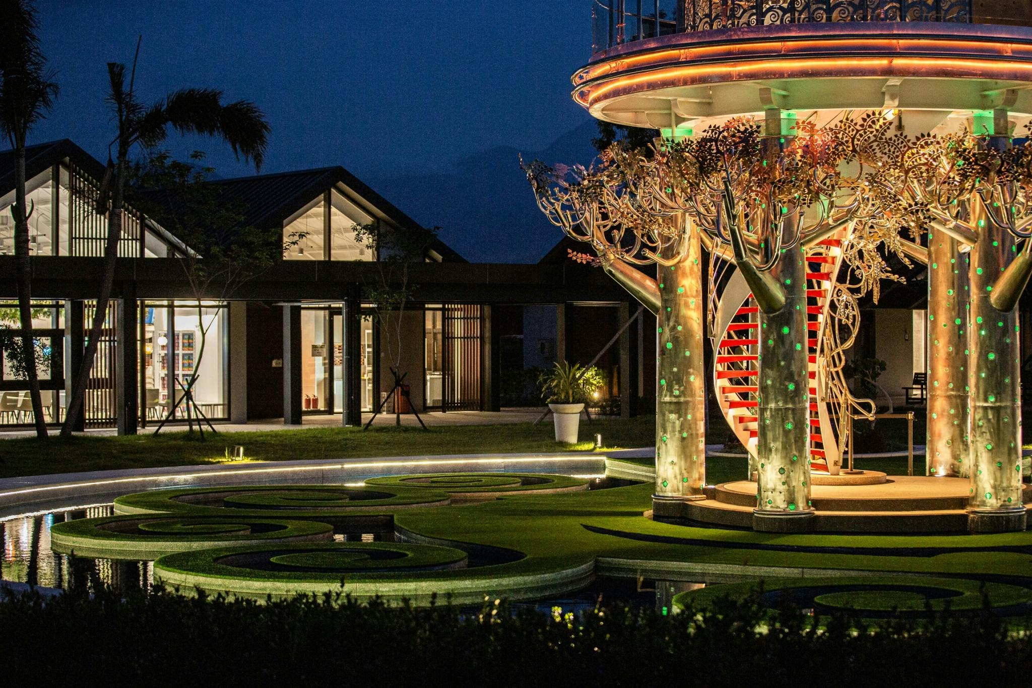 A decorative structure with illuminated details stands in a landscaped area at night, with a building in the background.
