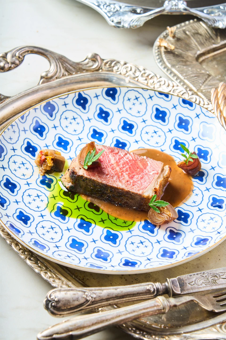A plated dish featuring a slice of cooked meat with sauce, garnishes, and decorative onions on a patterned plate.