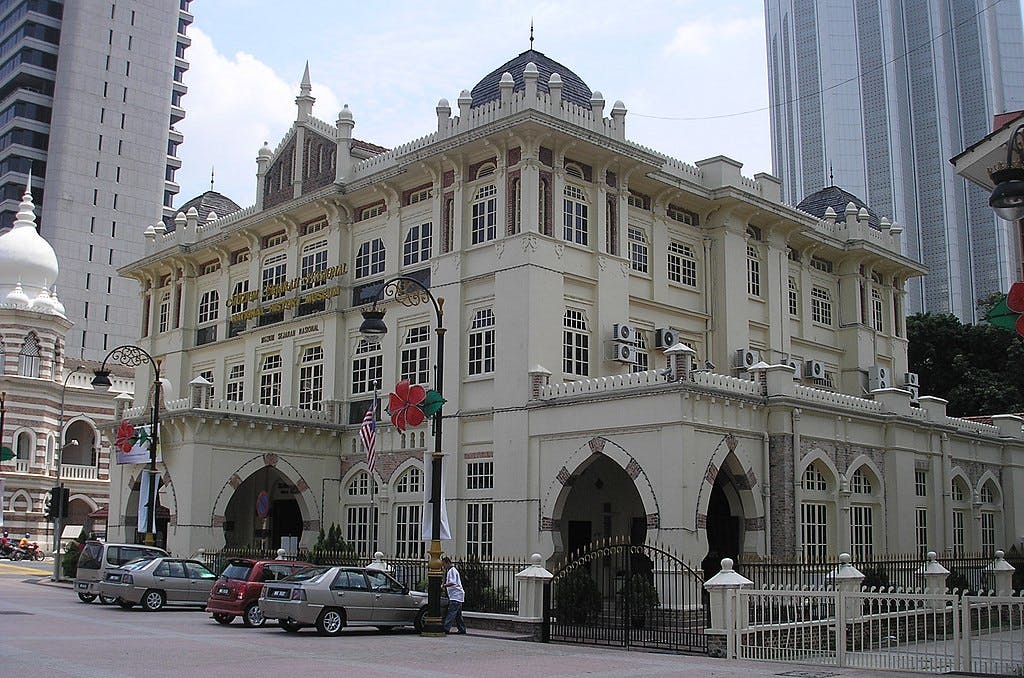 Historická béžová budova s klenutými okny, vlajkami a zaparkovanými auty před domem. Vysoké moderní mrakodrapy v pozadí.