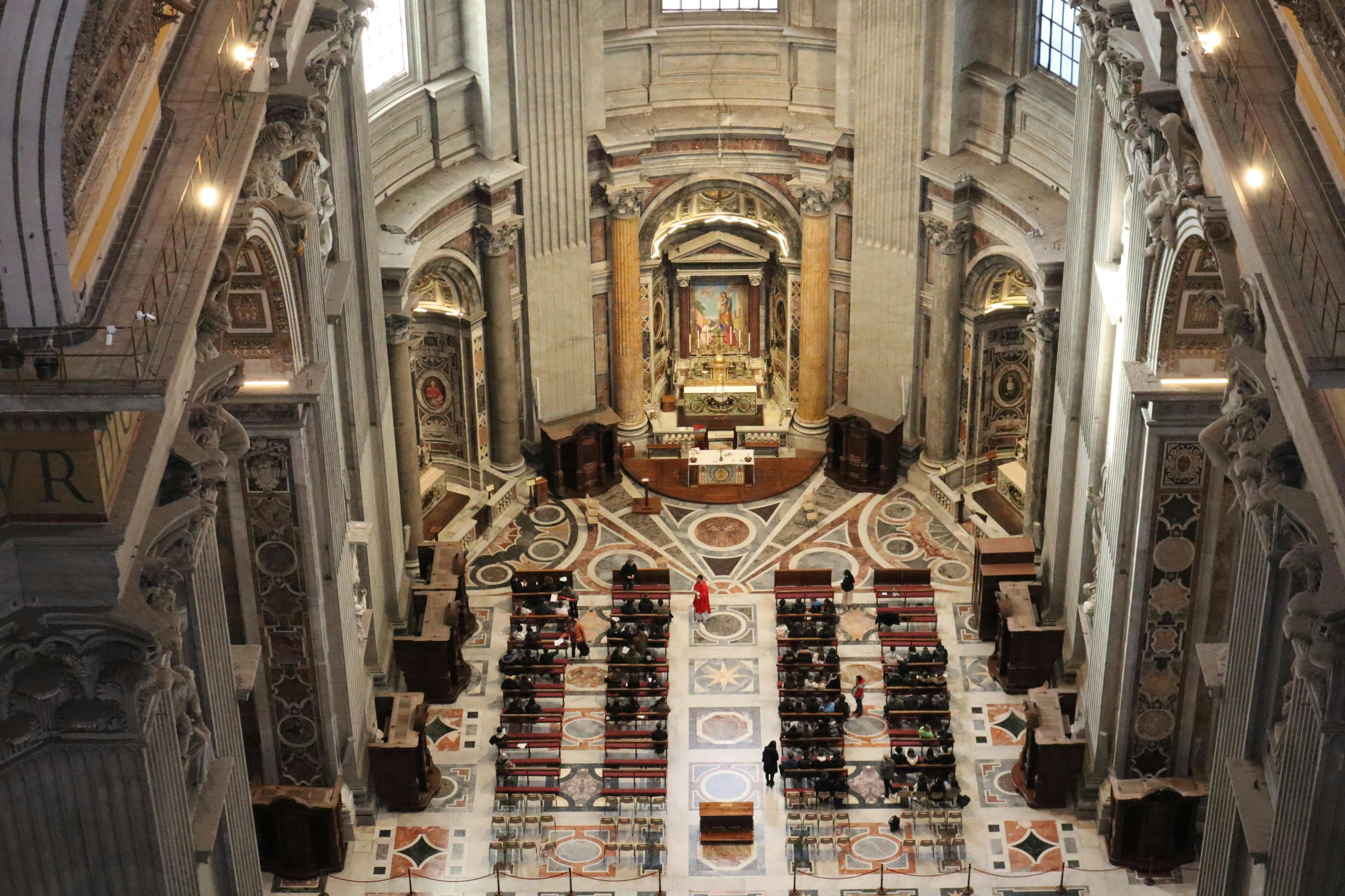 L&#39;interno di una grande chiesa con un altare decorato, alte colonne e file di banchi pieni di persone. Una persona vestita di rosso si trova nella navata.