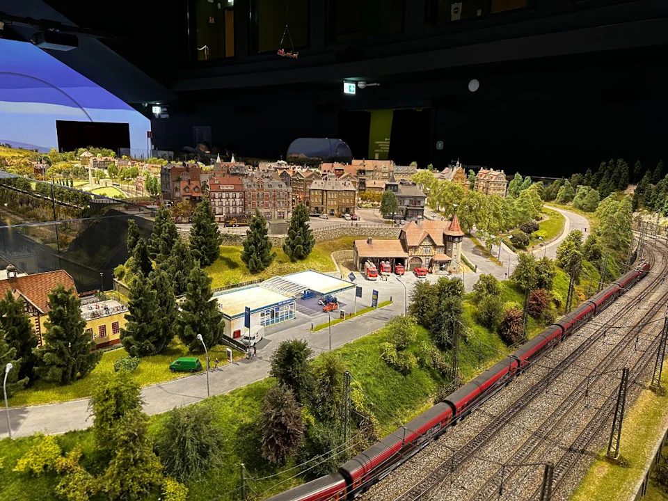 Detailed model train set featuring a town with buildings, trees, and cars. A red train runs along tracks in the foreground.