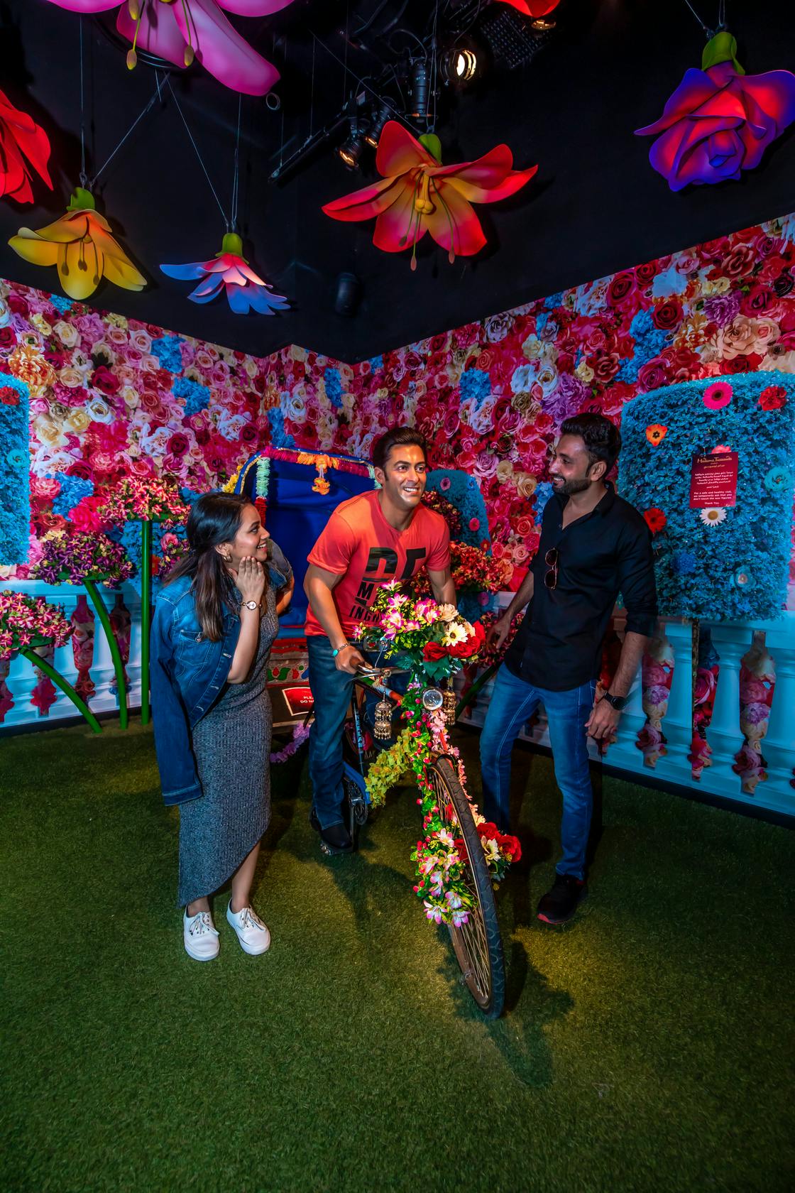 Tři lidé se pohybují kolem vyzdobeného kola v pulzující místnosti zdobené barevnými květinami a nadměrnými květinovými světly.